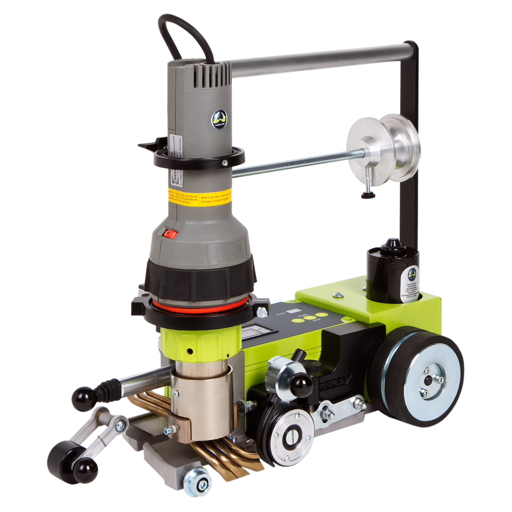 WOLFF_Dummy_56255_Weldmaster front_2017-07 Close-up of a compact industrial handheld machine mounted on a green metal base. The unit features a grey cylindrical motor housing with a black handle and ventilation slots, a red on/off rocker switch, and a yellow warning label wrapped around the upper body. A circular metal ring and a horizontal steel bar with an adjustable pulley assembly extend from the top, while the lower section shows precision adjustment knobs, polished metal rollers, drive wheels and several copper tubes or heating elements. The device combines plastic and metal components with visible fasteners and is designed for controlled material processing or pipe/joint work in a workshop setting.